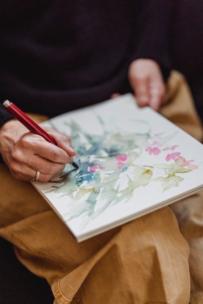 Close-up of hands painting delicate watercolor flowers and foliage in a sketchbook.