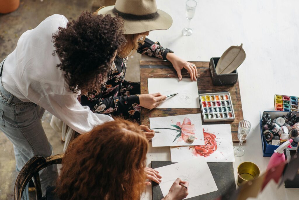 Women artists collaborating on watercolor flower paintings in an artistic studio setup.